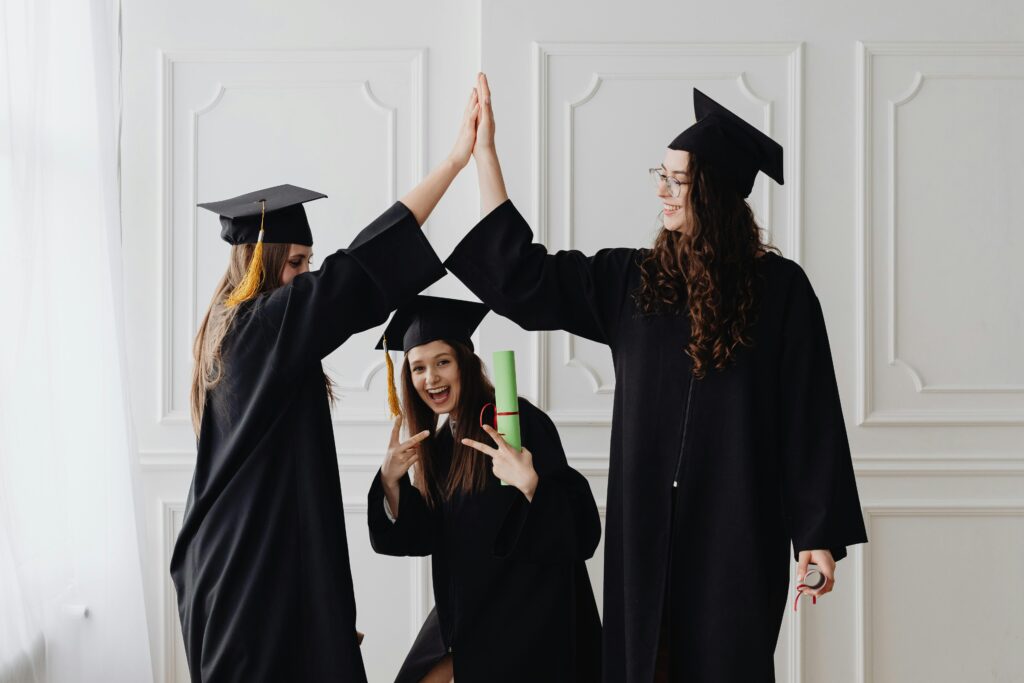 Three young women celebrating graduation with a high five, holding diplomas indoors.